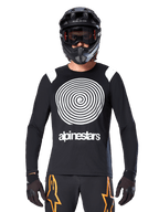 Camisola de Manga Comprida Alpinestars A-Supra Oscar, preta com gráfico em espiral branco e detalhes nos ombros, usada por um ciclista com capacete integral preto e óculos de proteção, calças pretas e cor-de-laranja, equipamento de ciclismo de performance para MTB gravity
