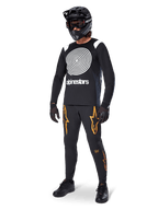Ciclista de MTB com camisola de manga comprida Alpinestars A-Supra Oscar em preto, com grafismo espiral vintage Oscar em branco, calças pretas com logótipos de estrela em cor de laranja, capacete integral preto com óculos e luvas pretas, equipamento profissional de BTT