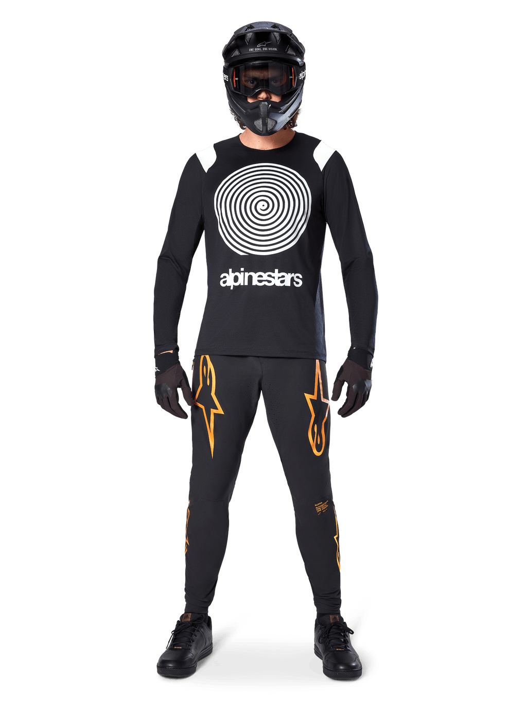 Modelo masculino com equipamento completo de mountain bike, Camisola de Manga Comprida Alpinestars A-Supra Oscar em preto com gráfico espiral branco no peito, calças pretas com logótipos de estrela cor de laranja, capacete integral e óculos pretos, luvas e sapatos pretos, conjunto coordenado para a prática de MTB gravity