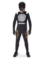 Modelo masculino com equipamento completo de mountain bike, Camisola de Manga Comprida Alpinestars A-Supra Oscar em preto com gráfico espiral branco no peito, calças pretas com logótipos de estrela cor de laranja, capacete integral e óculos pretos, luvas e sapatos pretos, conjunto coordenado para a prática de MTB gravity