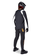 Camisola Alpinestars A-Supra Oscar de Manga Longa, preta com painéis brancos nas mangas, usada por um ciclista de MTB visto de costas, com capacete integral preto e calças pretas com logótipos cor de laranja, vestuário de ciclismo para gravidade off-road
