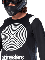 Camisola Alpinestars A-Supra Oscar, de manga comprida, preta, com um grande gráfico em espiral Oscar branco no peito e o logótipo da marca, usada por um ciclista com capacete integral preto e óculos de proteção, vestuário de BTT de performance para modalidades de gravidade