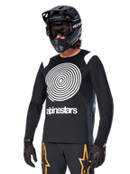 Alpinestars A-Supra Oscar Jersey de Manga Comprida, jersey de ciclismo MTB, preto e branco, com um design de espiral branca arrojado e logótipo Alpinestars no peito, usada com capacete integral preto e óculos de proteção, vestuário de performance para BTT
