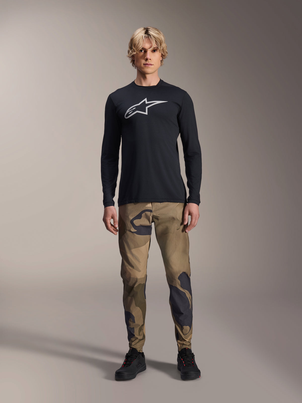 Alpinestars A-Dura Camo Pant, calças de ciclismo MTB, camuflagem castanha e verde, combinadas com camisola de manga comprida preta e sapatos pretos, estilo desportivo de mountain bike com tecido elástico durável.