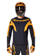 Ciclista de BTT equipado com camisola Alpinestars A-Supra Race de manga comprida, preta com gráficos cor de laranja e logótipo no peito, acompanhada por capacete integral laranja, óculos de proteção pretos e luvas cor de laranja, equipamento de competição coordenado para performance em BTT