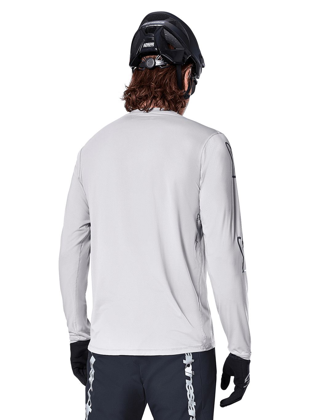 Camisola de Manga Longa Alpinestars A-Dura Switch, Cinzento Claro, vista traseira de um ciclista de montanha com capacete de BTT preto e luvas, destacando a cauda mais longa para maior cobertura e o tecido jacquard texturizado, equipamento de ciclismo de alta performance.