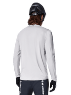 Camisola de Manga Longa Alpinestars A-Dura Switch, Cinzento Claro, vista traseira de um ciclista de montanha com capacete de BTT preto e luvas, destacando a cauda mais longa para maior cobertura e o tecido jacquard texturizado, equipamento de ciclismo de alta performance.