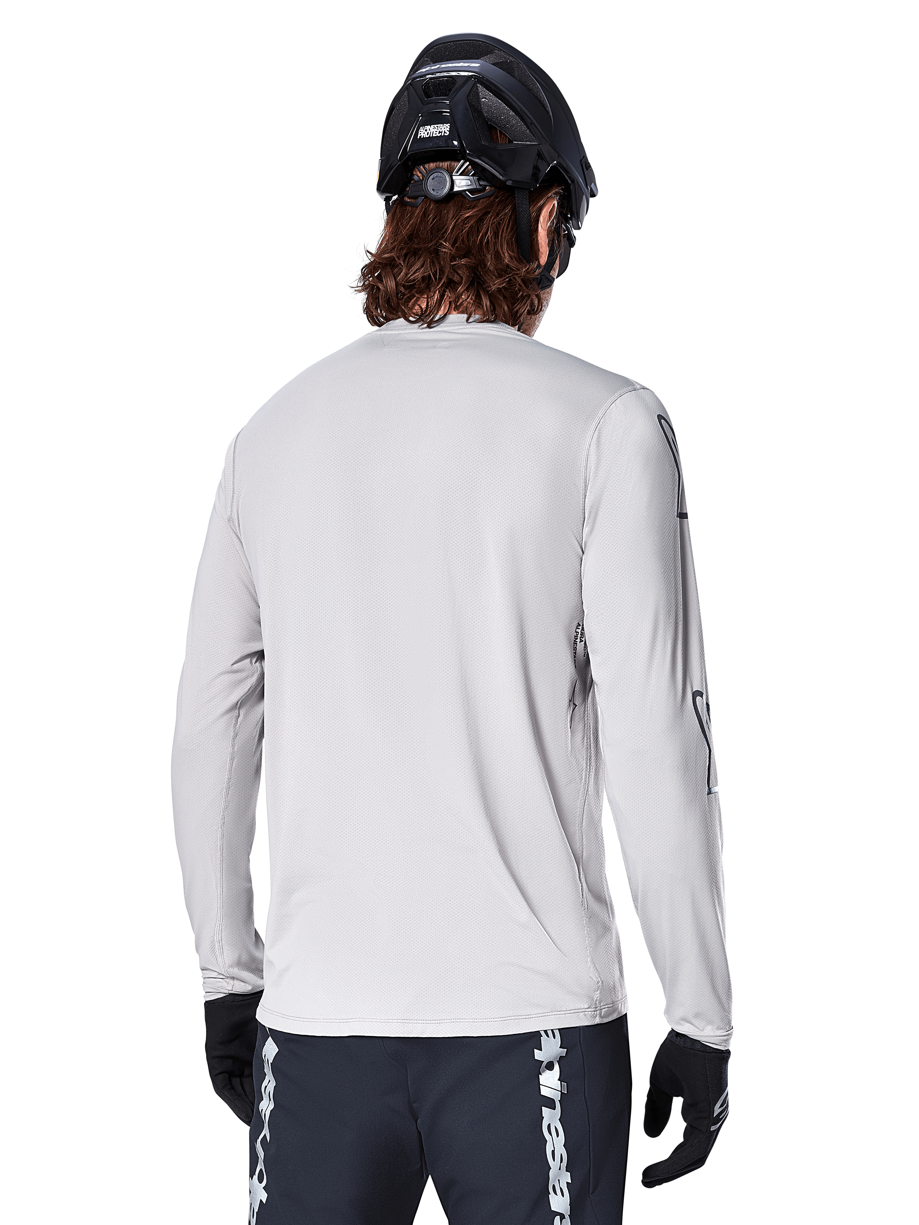Camisola de Manga Longa Alpinestars A-Dura Switch, Cinzento Claro, vista traseira de um ciclista de montanha com capacete de BTT preto e luvas, destacando a cauda mais longa para maior cobertura e o tecido jacquard texturizado, equipamento de ciclismo de alta performance.