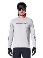 Camisola de Manga Comprida Alpinestars A-Dura Switch, Cinzento Claro, apresenta logótipo nominativo no peito e logótipos de estrela nas mangas em cinzento escuro, usada por um ciclista com capacete de BTT e luvas pretas, vestuário de ciclismo de performance para trilhos