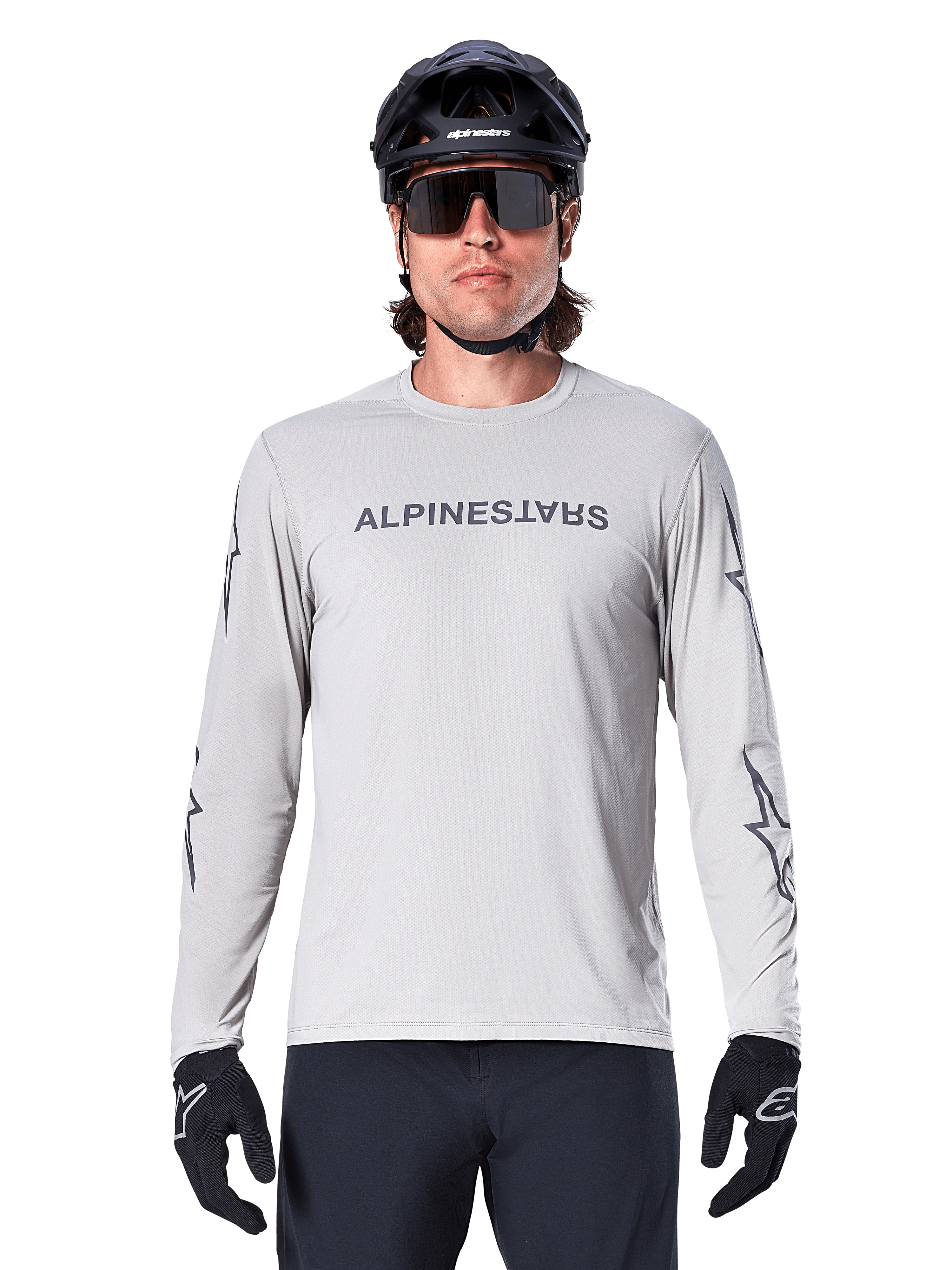 Camisola de Manga Comprida Alpinestars A-Dura Switch, Cinzento Claro, apresenta logótipo nominativo no peito e logótipos de estrela nas mangas em cinzento escuro, usada por um ciclista com capacete de BTT e luvas pretas, vestuário de ciclismo de performance para trilhos