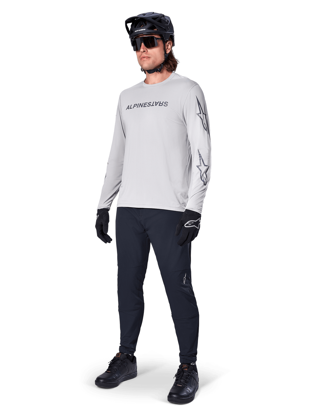 Camisola de Manga Comprida Alpinestars A-Dura Switch, cinzento claro, usada por ciclista masculino com calças de BTT pretas e capacete, com logótipo nominativo preto no peito e grafismos de estrela nas mangas, equipamento de ciclismo de alta performance para todo-o-terreno