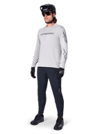 Camisola de Manga Comprida Alpinestars A-Dura Switch, cinzento claro, usada por ciclista masculino com calças de BTT pretas e capacete, com logótipo nominativo preto no peito e grafismos de estrela nas mangas, equipamento de ciclismo de alta performance para todo-o-terreno