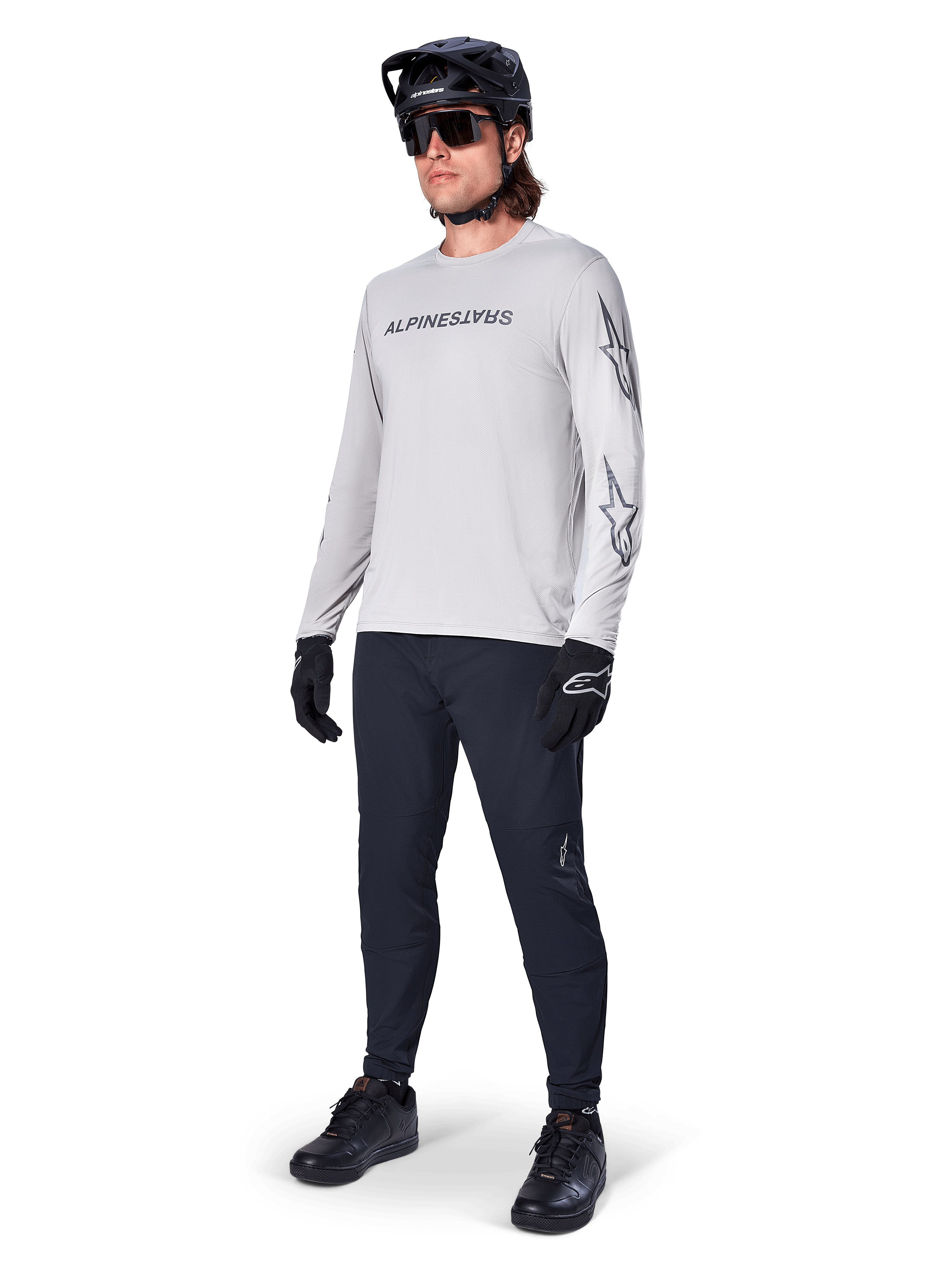 Camisola de Manga Comprida Alpinestars A-Dura Switch, cinzento claro, usada por ciclista masculino com calças de BTT pretas e capacete, com logótipo nominativo preto no peito e grafismos de estrela nas mangas, equipamento de ciclismo de alta performance para todo-o-terreno