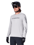 Alpinestars A-Dura Switch Long Sleeve Jersey, camisola de BTT, cinzento claro, logótipo nominativo em serigrafia preta no peito e logótipos de estrela nas mangas, usada com capacete de BTT preto, óculos de sol escuros e luvas pretas, equipamento atlético de ciclismo todo-o-terreno