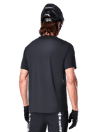 Camisola Alpinestars A-Dura Switch de Manga Curta, preta, vista traseira num modelo com capacete de ciclismo preto e calções de BTT, realçando o ajuste regular e a parte traseira mais comprida desenhada para ciclismo de montanha