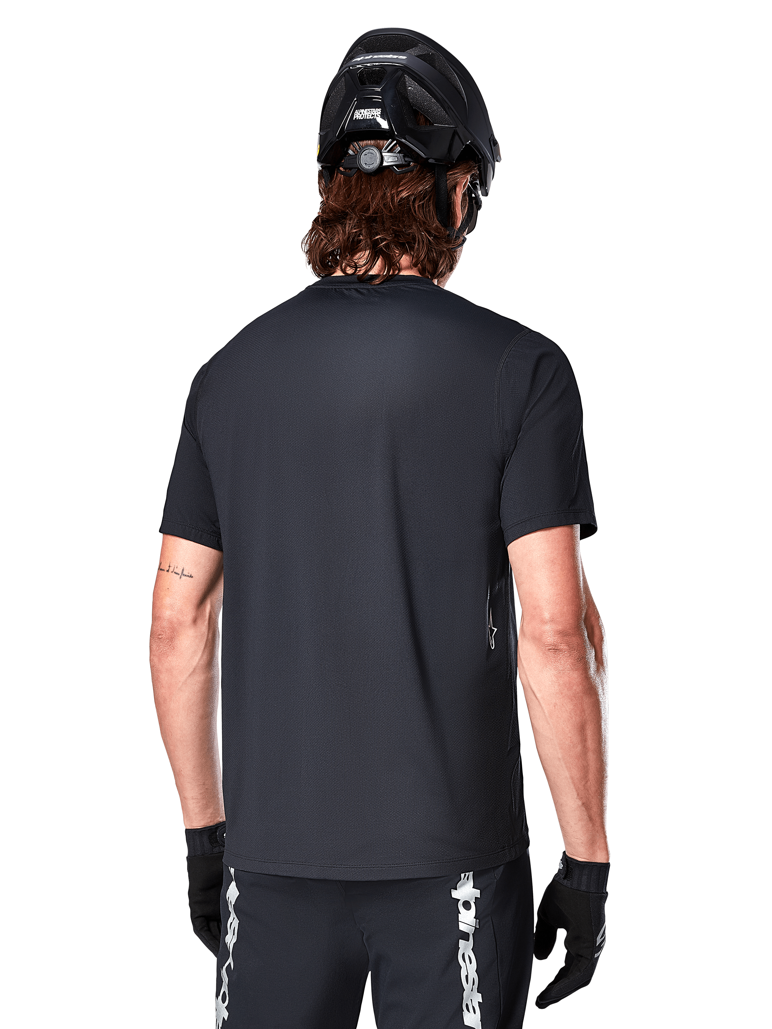 Camisola Alpinestars A-Dura Switch de Manga Curta, preta, vista traseira num modelo com capacete de ciclismo preto e calções de BTT, realçando o ajuste regular e a parte traseira mais comprida desenhada para ciclismo de montanha