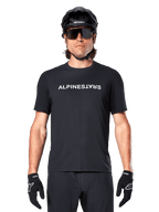 Ciclista de montanha a usar a Camisola de Manga Curta Alpinestars A-Dura Switch em preto, com um logótipo de texto branco no peito, combinada com um capacete preto, óculos de sol grandes e luvas pretas, vestuário de ciclismo profissional para trilhos.
