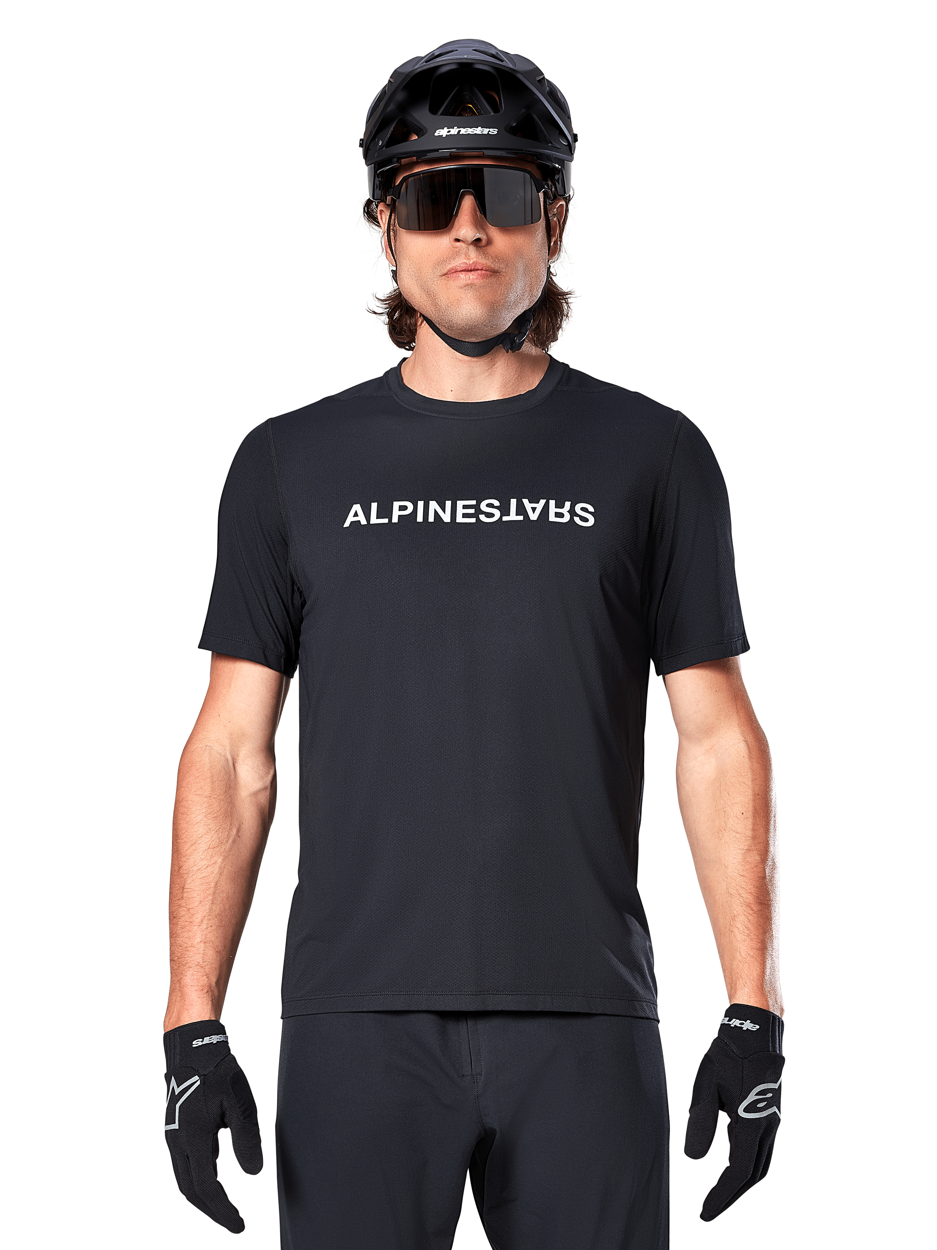 Ciclista de montanha a usar a Camisola de Manga Curta Alpinestars A-Dura Switch em preto, com um logótipo de texto branco no peito, combinada com um capacete preto, óculos de sol grandes e luvas pretas, vestuário de ciclismo profissional para trilhos.