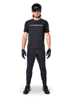Ciclista de BTT com a Camisola Alpinestars A-Dura Switch de Manga Curta em preto, com logótipo estilizado a branco no peito, calças de BTT pretas, capacete e luvas, equipamento completo de ciclismo pronto para trilhos.