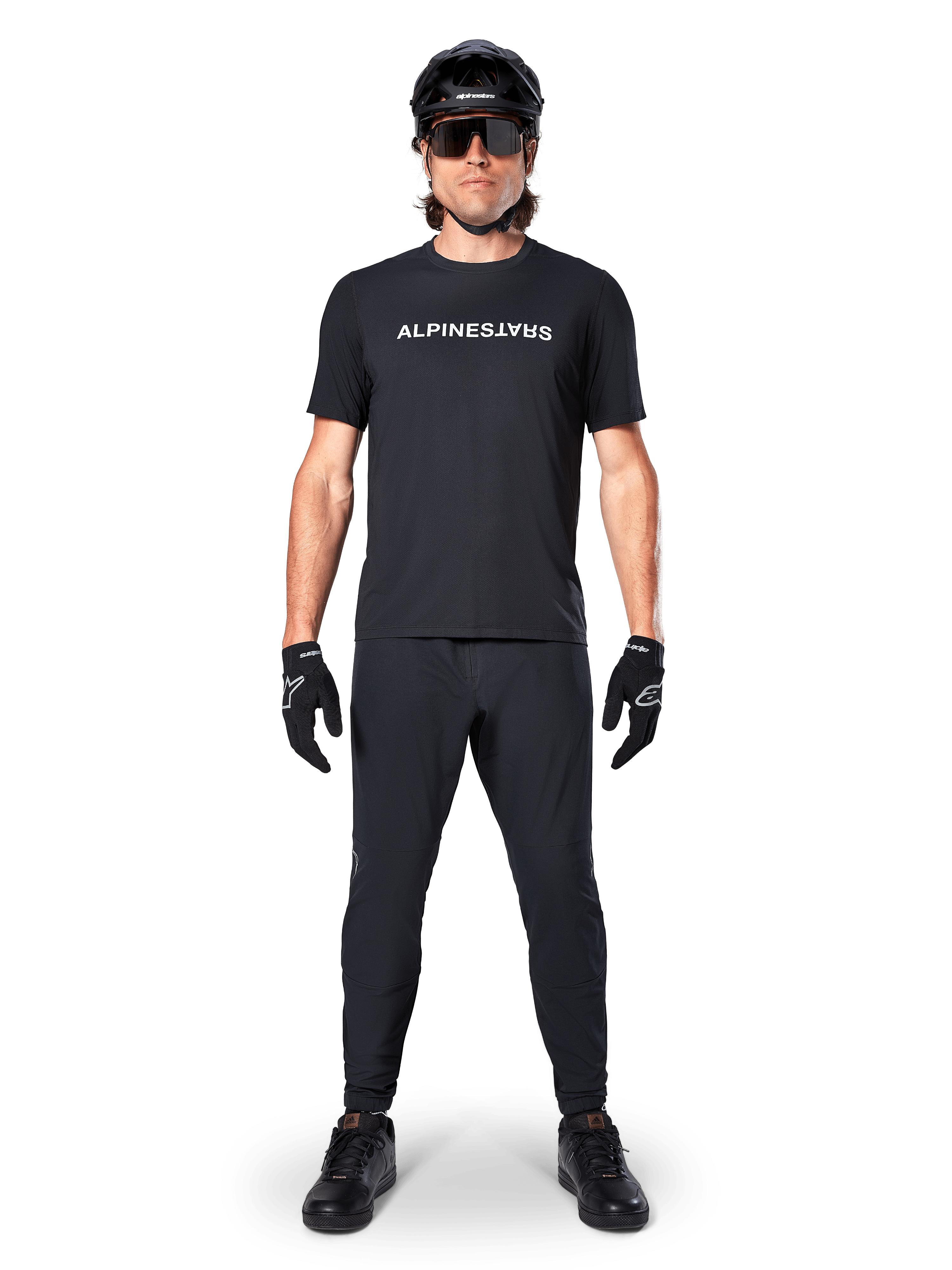 Ciclista de BTT com a Camisola Alpinestars A-Dura Switch de Manga Curta em preto, com logótipo estilizado a branco no peito, calças de BTT pretas, capacete e luvas, equipamento completo de ciclismo pronto para trilhos.