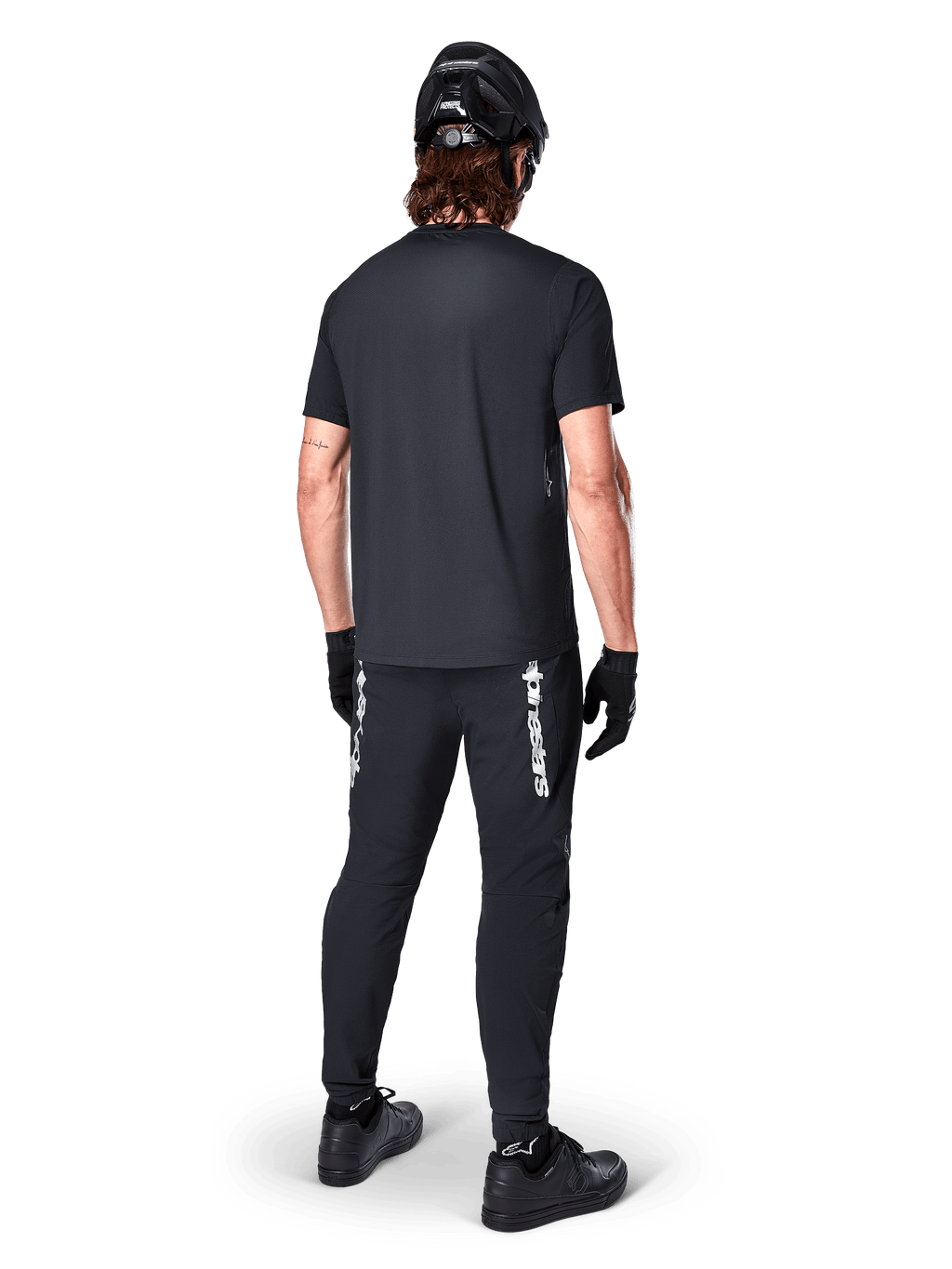 Alpinestars A-Dura Switch Jersey - Short Sleeve, preta, vista traseira de ciclista a usar equipamento completo de bicicleta de montanha, incluindo calças pretas a condizer com logótipo nominativo branco, capacete preto e luvas, vestuário de ciclismo de performance para trilhos