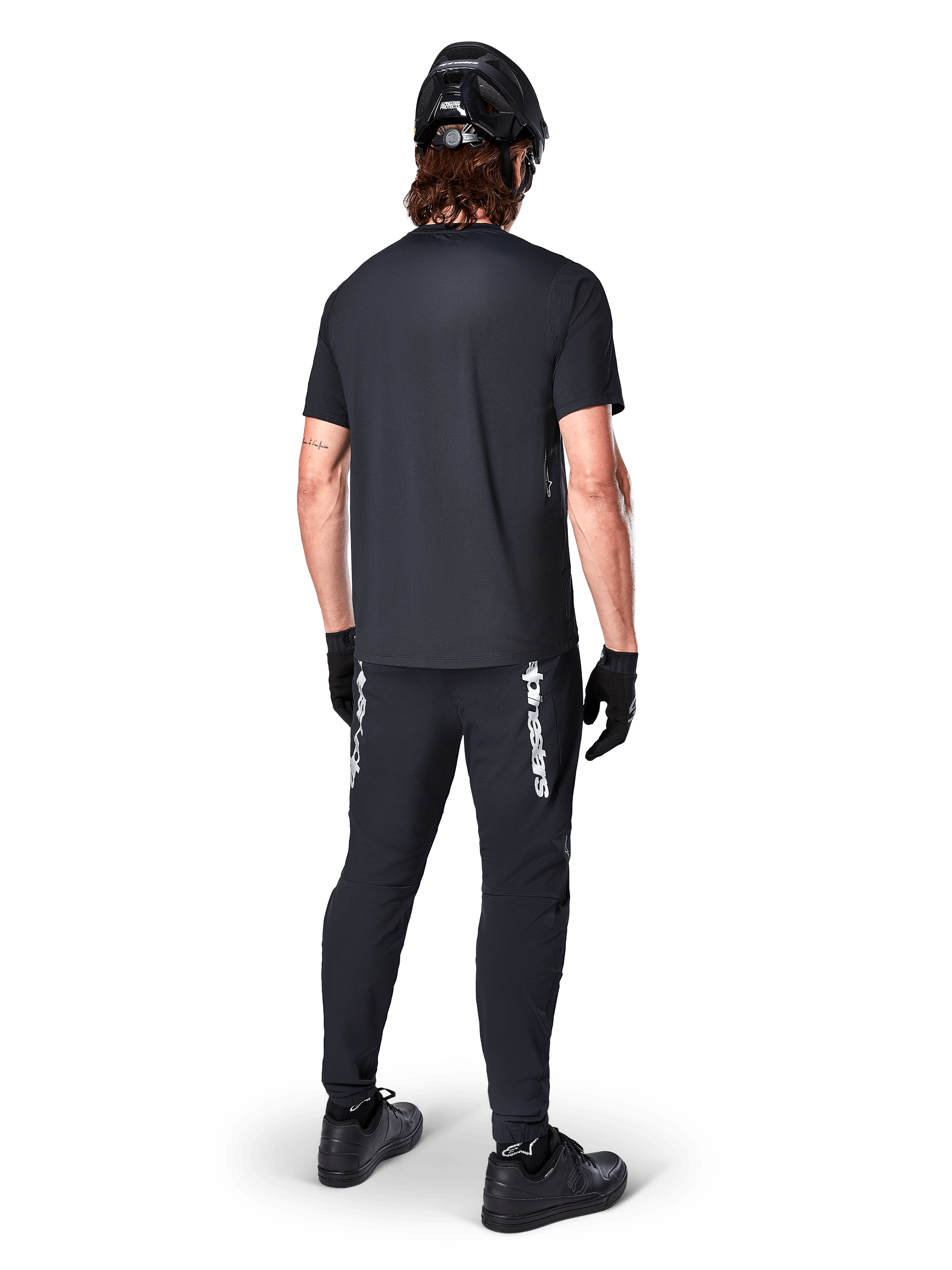 Alpinestars A-Dura Switch Jersey - Short Sleeve, preta, vista traseira de ciclista a usar equipamento completo de bicicleta de montanha, incluindo calças pretas a condizer com logótipo nominativo branco, capacete preto e luvas, vestuário de ciclismo de performance para trilhos