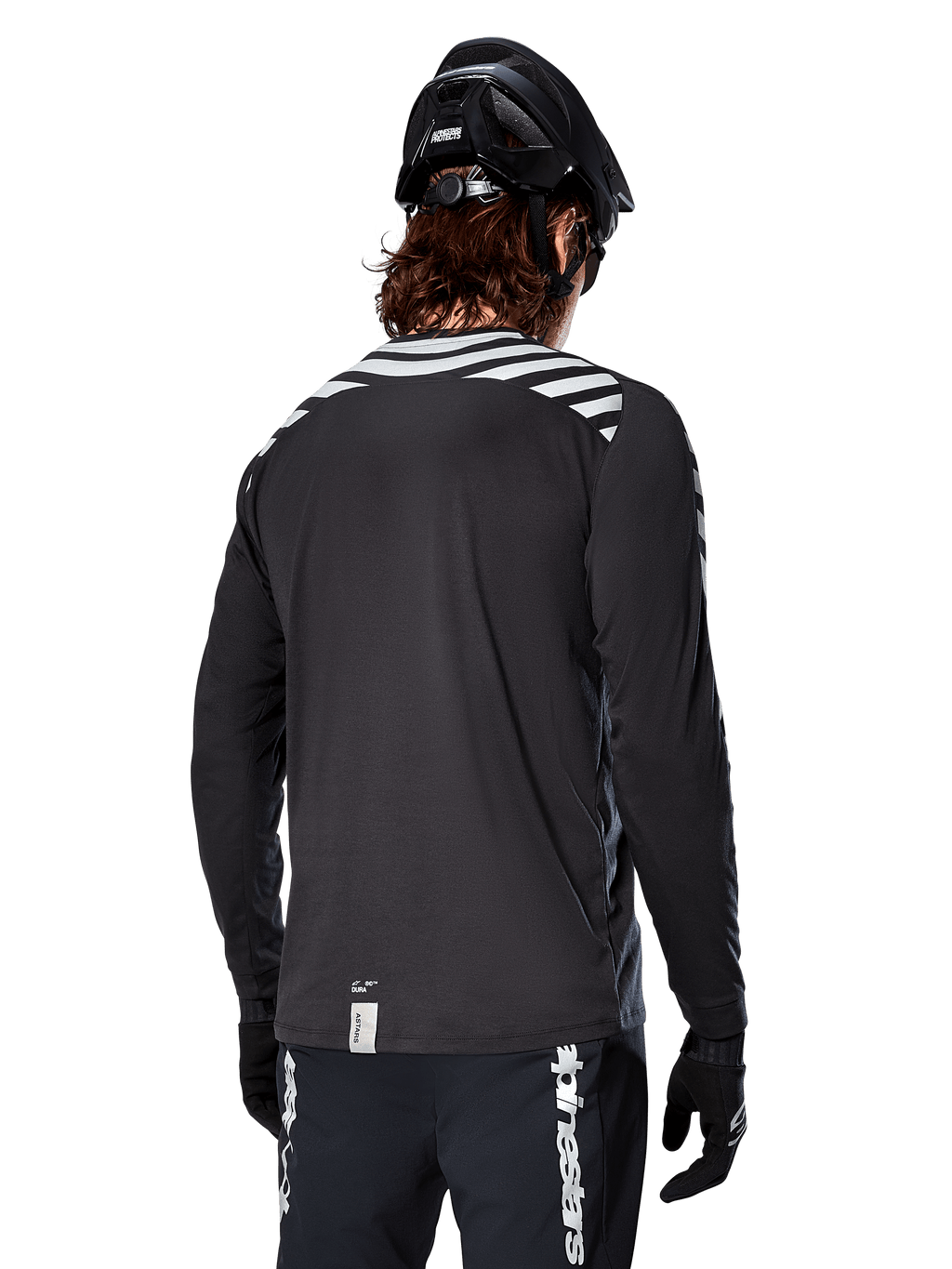 Camisola de Manga Comprida Alpinestars A-Dura Dri Oscar, preto refletor, vista traseira com riscas diagonais refletoras nos ombros e parte traseira mais comprida, usada com capacete e luvas de BTT pretos, equipamento técnico para BTT