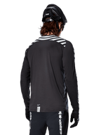 Camisola de Manga Comprida Alpinestars A-Dura Dri Oscar, preto refletor, vista traseira com riscas diagonais refletoras nos ombros e parte traseira mais comprida, usada com capacete e luvas de BTT pretos, equipamento técnico para BTT