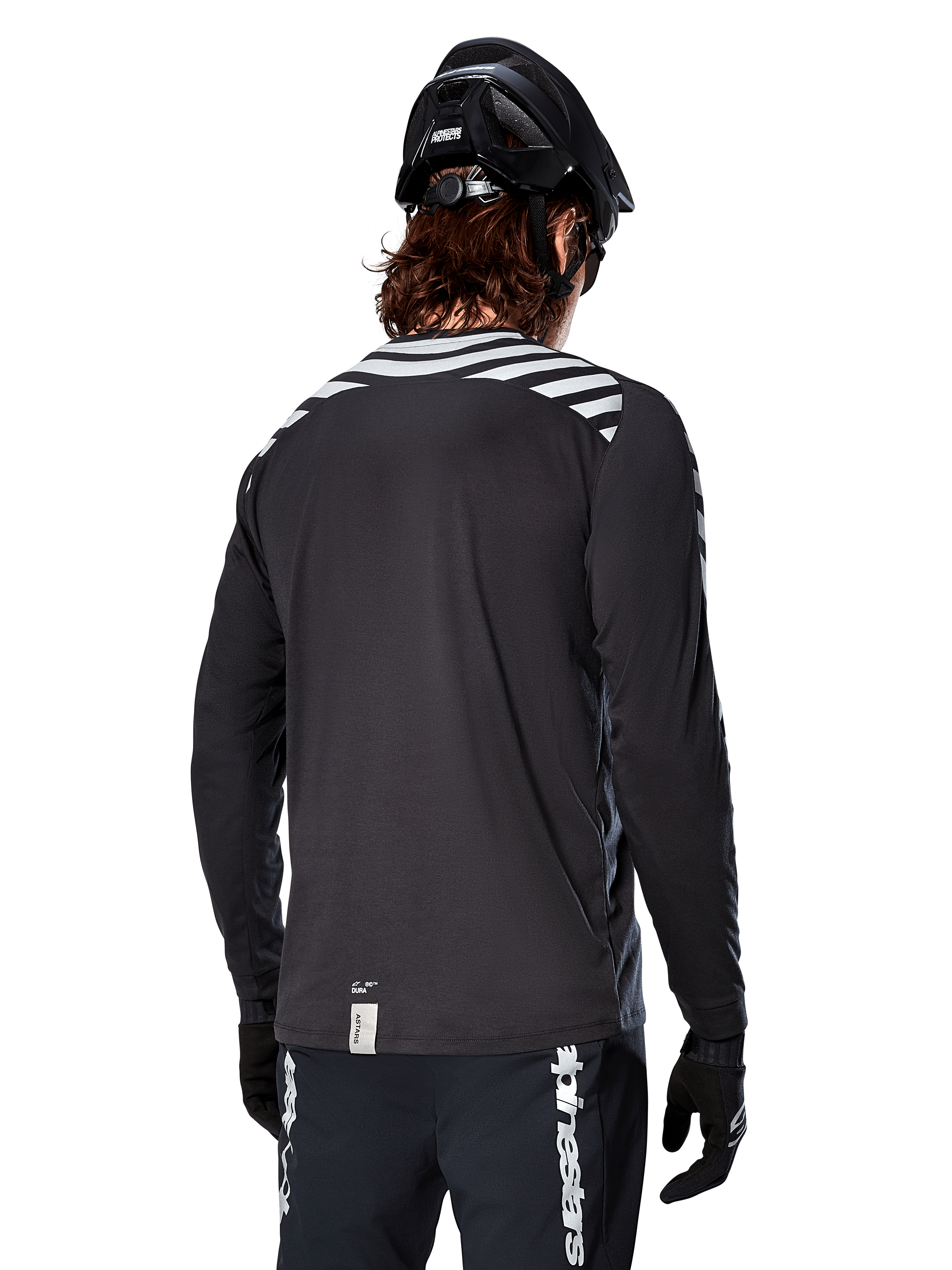 Camisola de Manga Comprida Alpinestars A-Dura Dri Oscar, preto refletor, vista traseira com riscas diagonais refletoras nos ombros e parte traseira mais comprida, usada com capacete e luvas de BTT pretos, equipamento técnico para BTT