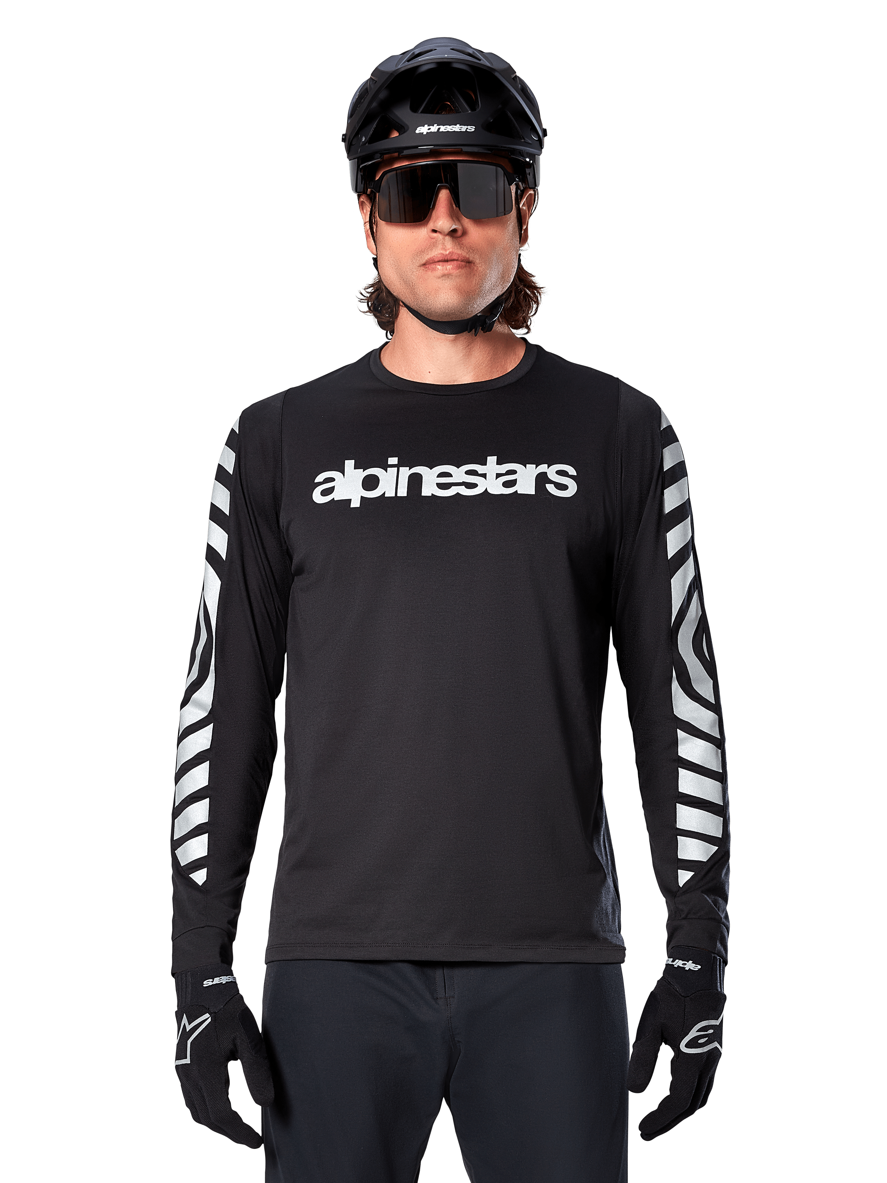 Ciclista masculino com Camisola Alpinestars A-Dura Dri Oscar de Manga Comprida, preto refletor, com logótipo Alpinestars branco no peito e gráficos prateados refletores nas mangas, acompanhado por capacete de BTT preto, óculos de sol e luvas pretas, equipamento de ciclismo de performance
