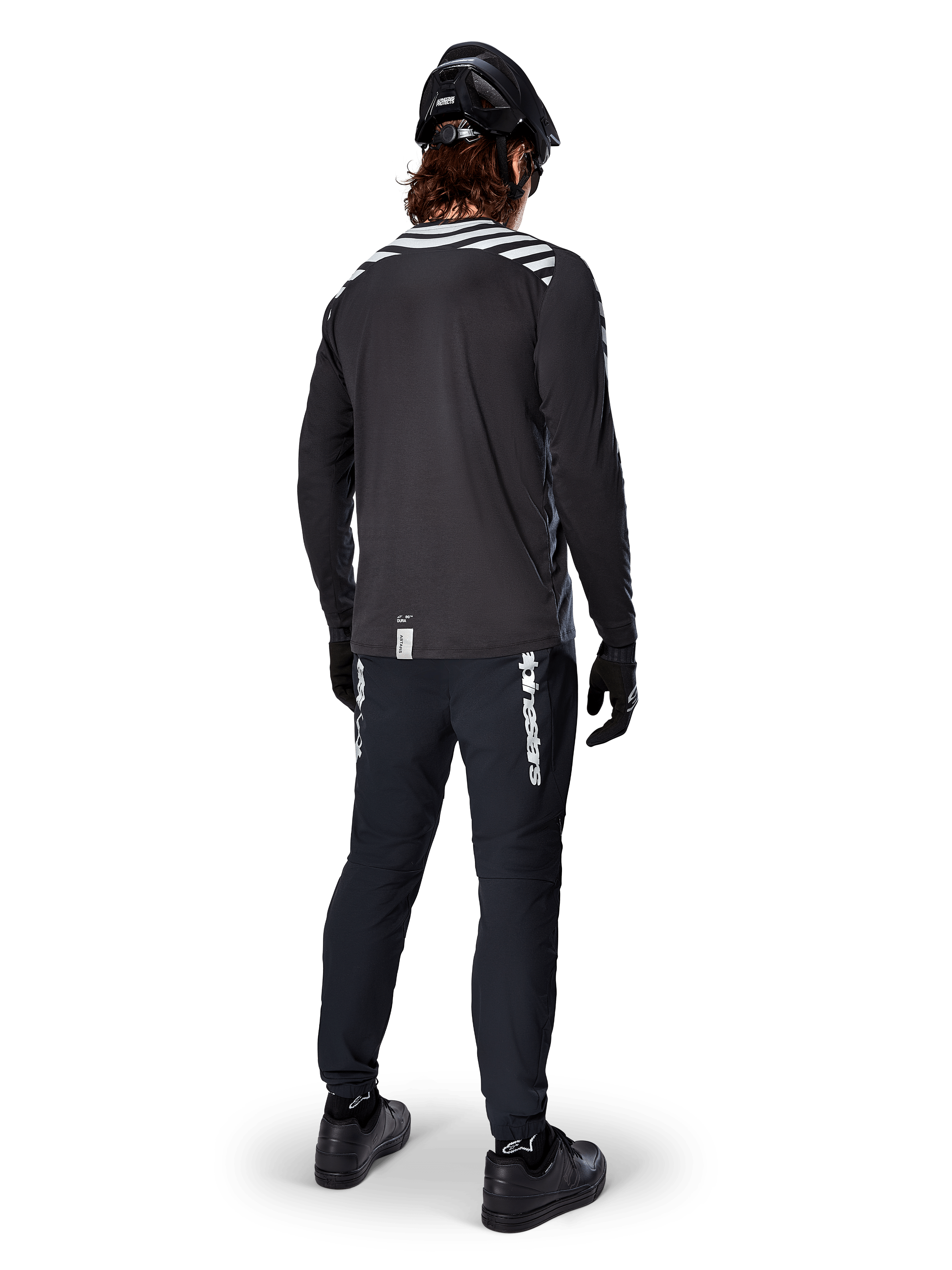 Ciclista de BTT com a camisola de manga comprida Alpinestars A-Dura Dri Oscar em Preto Refletor, vista de costas, calças técnicas pretas, capacete e luvas, com riscas refletoras nos ombros e cauda mais longa para ciclismo todo-o-terreno.