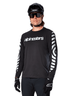 Camisola de manga comprida Alpinestars A-Dura Dri Oscar, camisola de ciclismo MTB, preto refletor, com logótipo branco no peito e gráficos circulares refletivos arrojados nas mangas, utilizada com um capacete preto e óculos de sol, vestuário técnico com gestão de humidade para ciclismo de montanha fora de estrada
