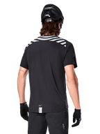 Alpinestars A-Dura Dri Oscar Jersey, camisola de MTB de manga curta, preto refletor, vista traseira num modelo a usar capacete de ciclismo e luvas, com riscas cinzentas refletoras nos ombros e cauda mais longa, tecido técnico com gestão de humidade para mountain bike