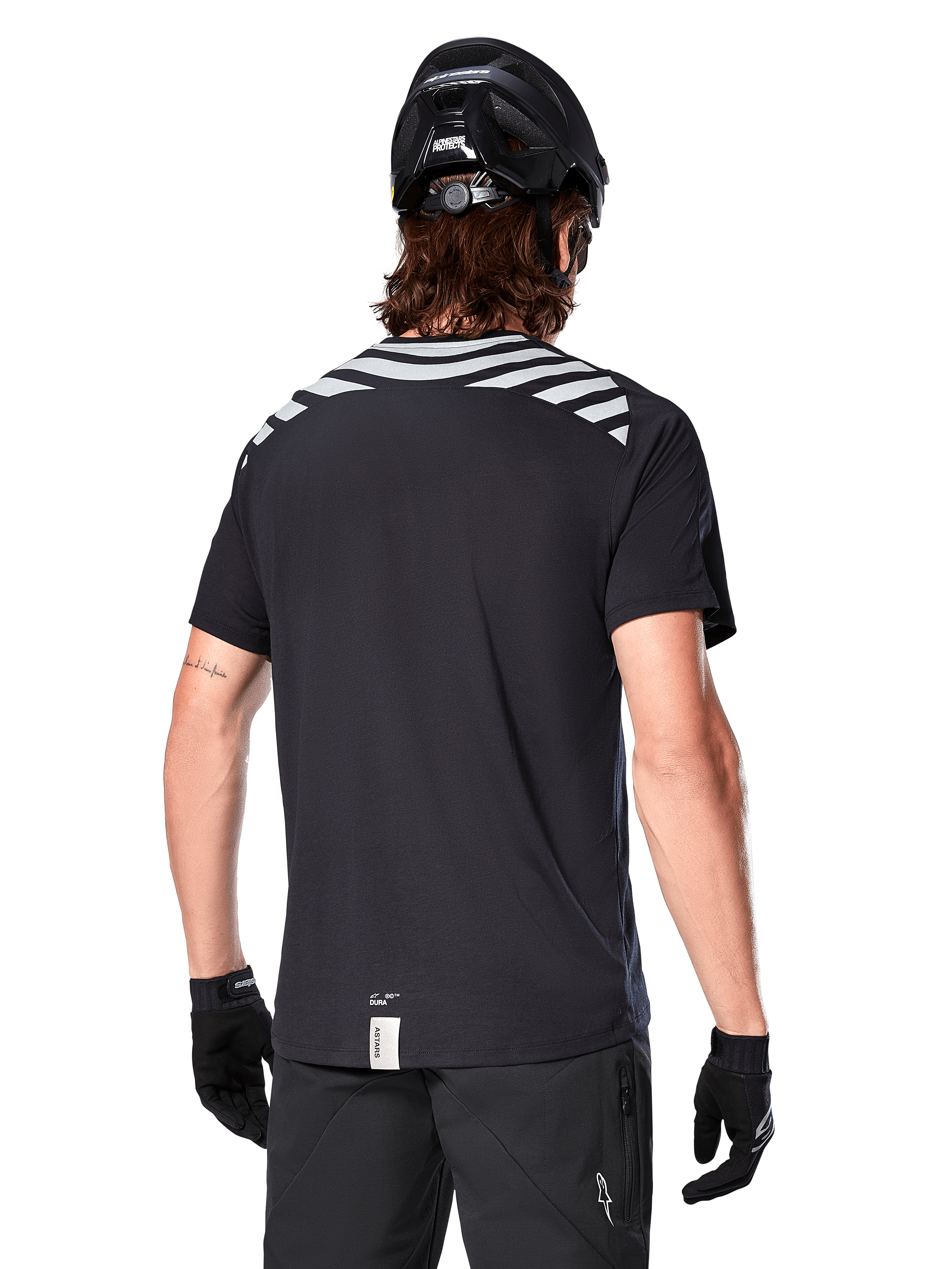 Alpinestars A-Dura Dri Oscar Jersey, camisola de MTB de manga curta, preto refletor, vista traseira num modelo a usar capacete de ciclismo e luvas, com riscas cinzentas refletoras nos ombros e cauda mais longa, tecido técnico com gestão de humidade para mountain bike