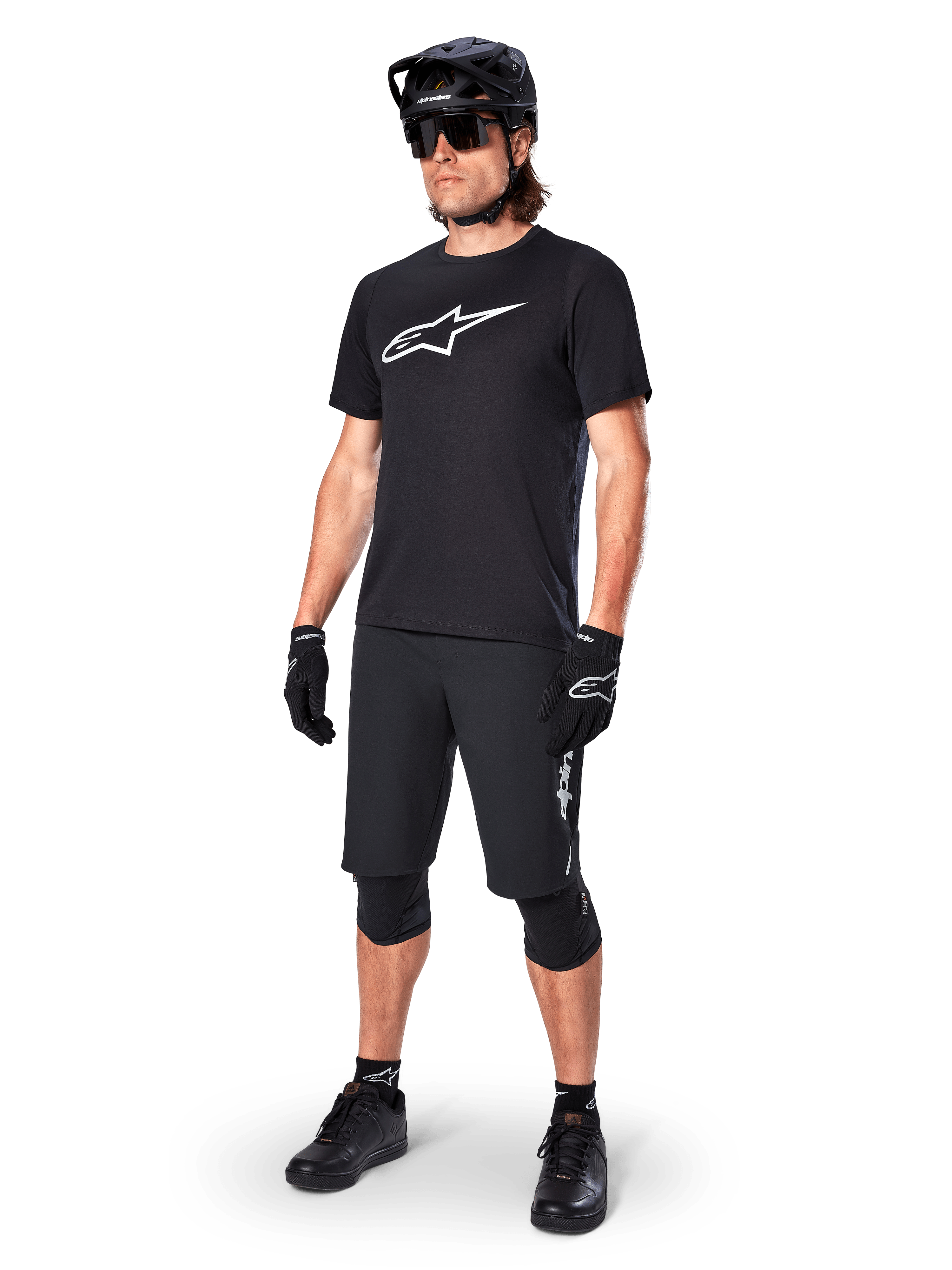 Ciclista de BTT a usar a camisola de manga curta Alpinestars A-Dura Dri Oscar em preto refletor, com um logótipo branco de grandes dimensões no peito, combinada com calções de BTT pretos, luvas e capacete, vestuário técnico de ciclismo para performance fora de estrada