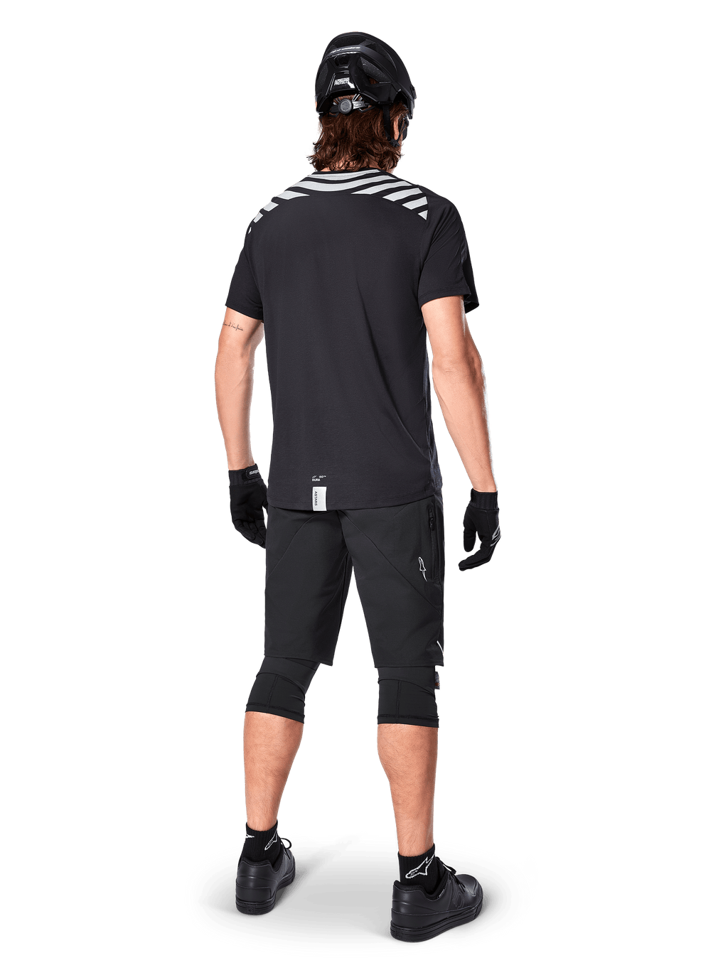 Alpinestars A-Dura Dri Oscar Short Sleeve Jersey, Preto Refletor, vista traseira de um ciclista a usar a camisola com calções de BTT pretos, capacete e joelheiras, com grafismos refletores nos ombros e design de cauda longa para a prática de BTT.