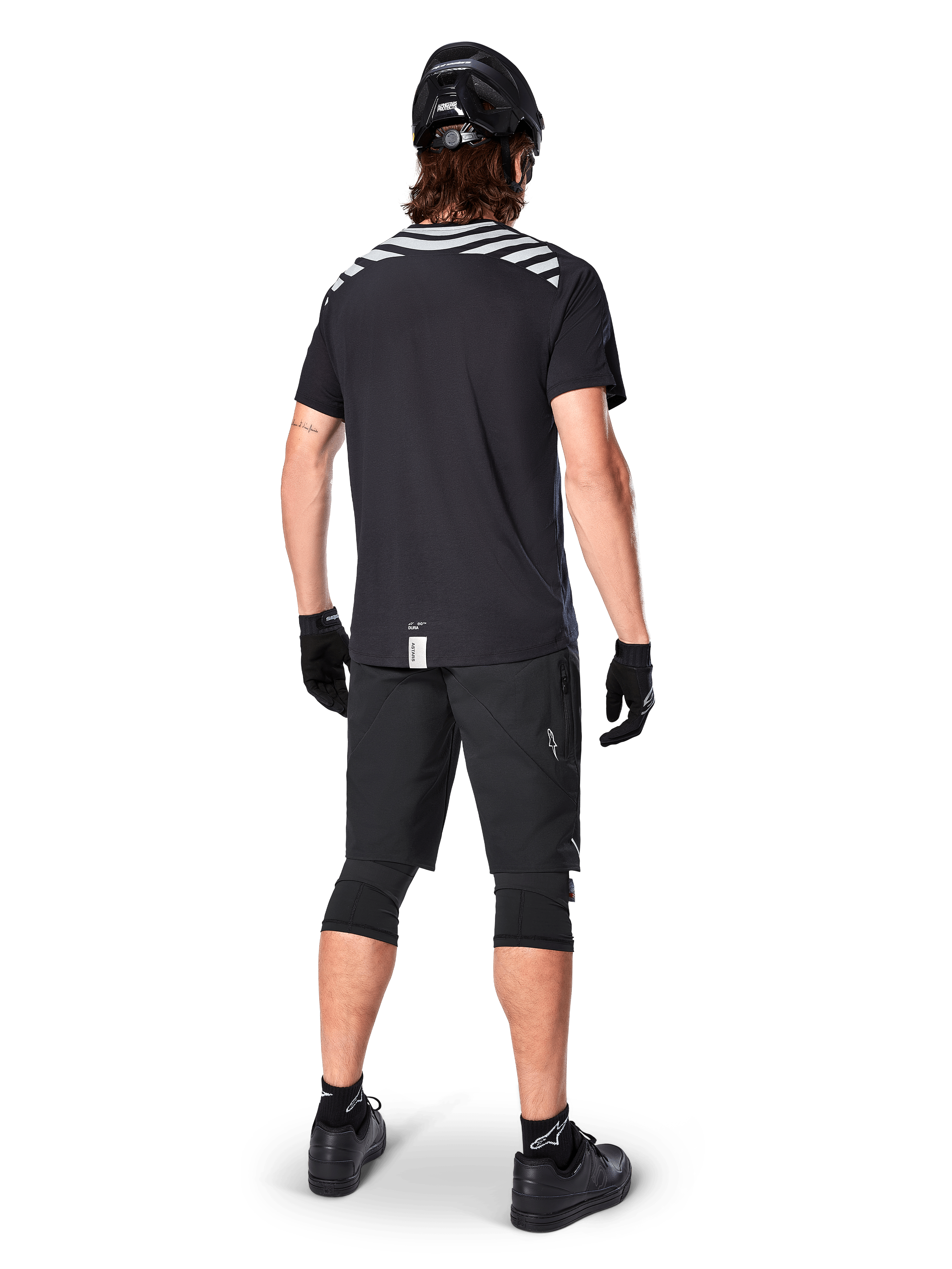 Alpinestars A-Dura Dri Oscar Short Sleeve Jersey, Preto Refletor, vista traseira de um ciclista a usar a camisola com calções de BTT pretos, capacete e joelheiras, com grafismos refletores nos ombros e design de cauda longa para a prática de BTT.
