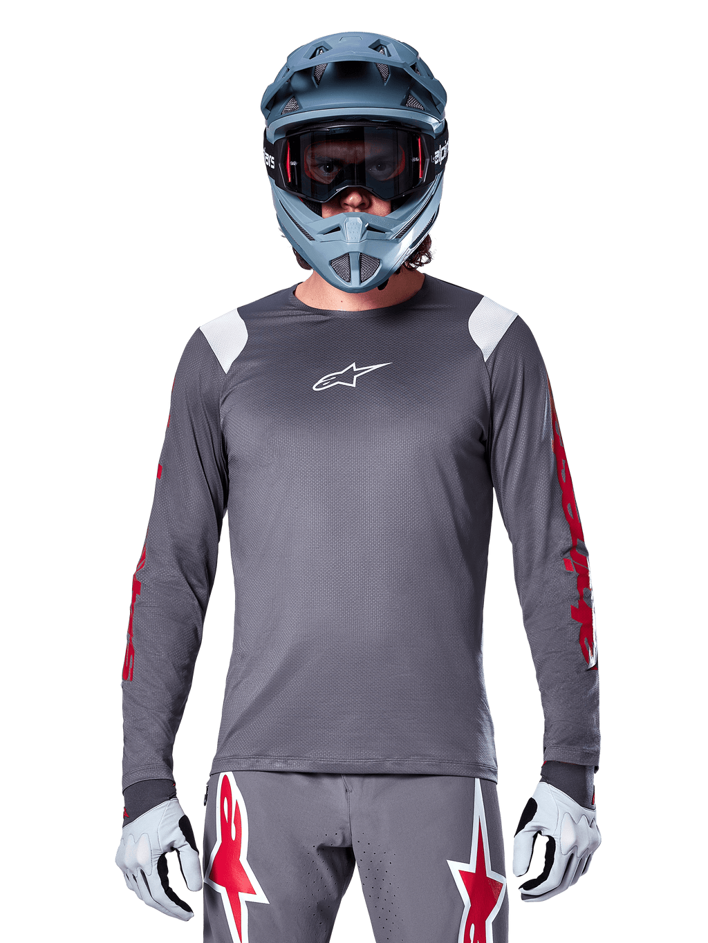 Ciclista de BTT a usar camisola de manga longa Alpinestars A-Supra Astar, cinzento escuro com grafismos vermelhos nas mangas e detalhes brancos nos ombros, calças cinzentas a condizer com logótipos de estrela vermelhos, capacete integral azul e óculos de proteção, equipamento de ciclismo todo-o-terreno coordenado