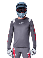 Ciclista de BTT a usar camisola de manga longa Alpinestars A-Supra Astar, cinzento escuro com grafismos vermelhos nas mangas e detalhes brancos nos ombros, calças cinzentas a condizer com logótipos de estrela vermelhos, capacete integral azul e óculos de proteção, equipamento de ciclismo todo-o-terreno coordenado