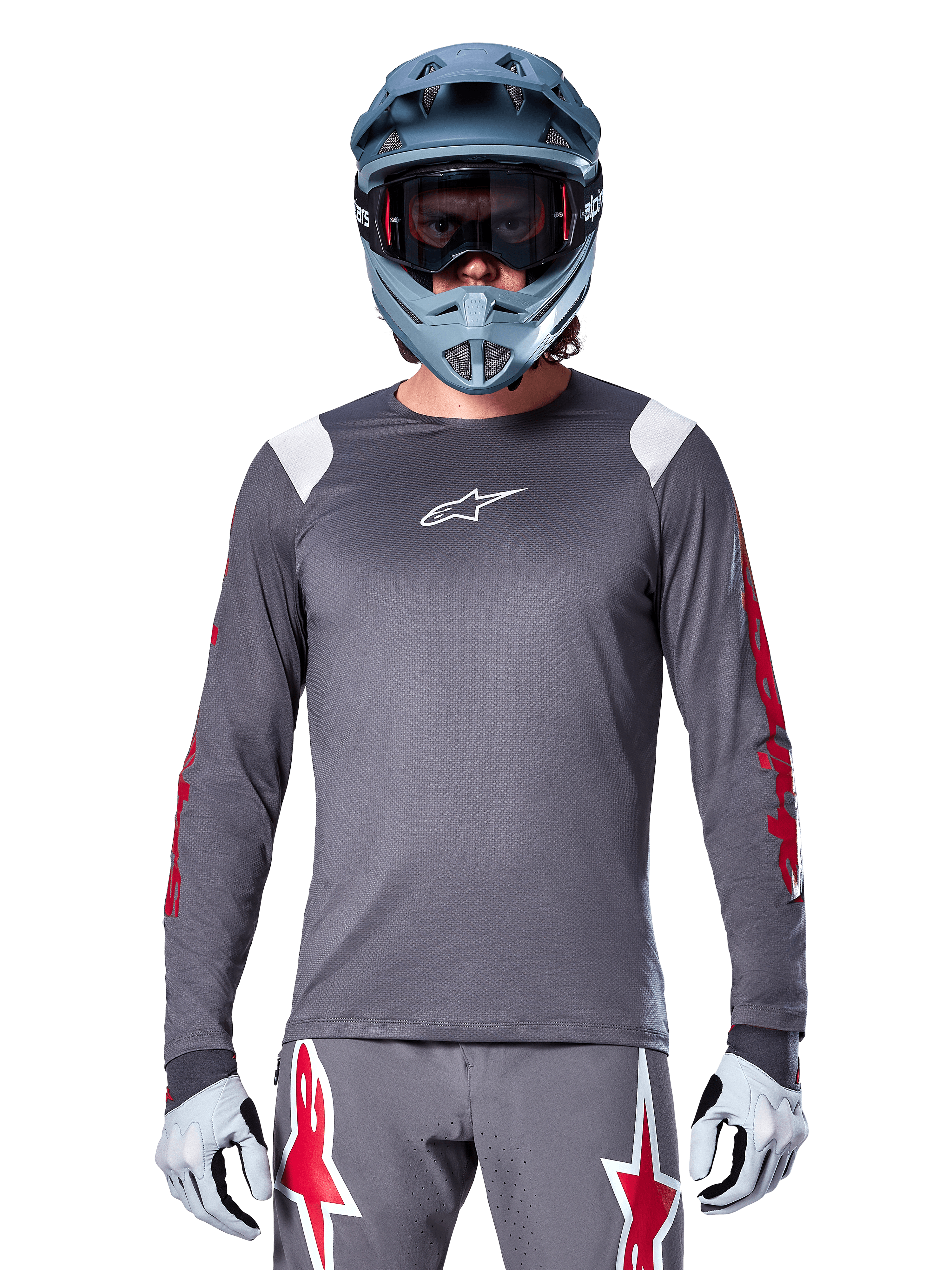 Ciclista de BTT a usar camisola de manga longa Alpinestars A-Supra Astar, cinzento escuro com grafismos vermelhos nas mangas e detalhes brancos nos ombros, calças cinzentas a condizer com logótipos de estrela vermelhos, capacete integral azul e óculos de proteção, equipamento de ciclismo todo-o-terreno coordenado