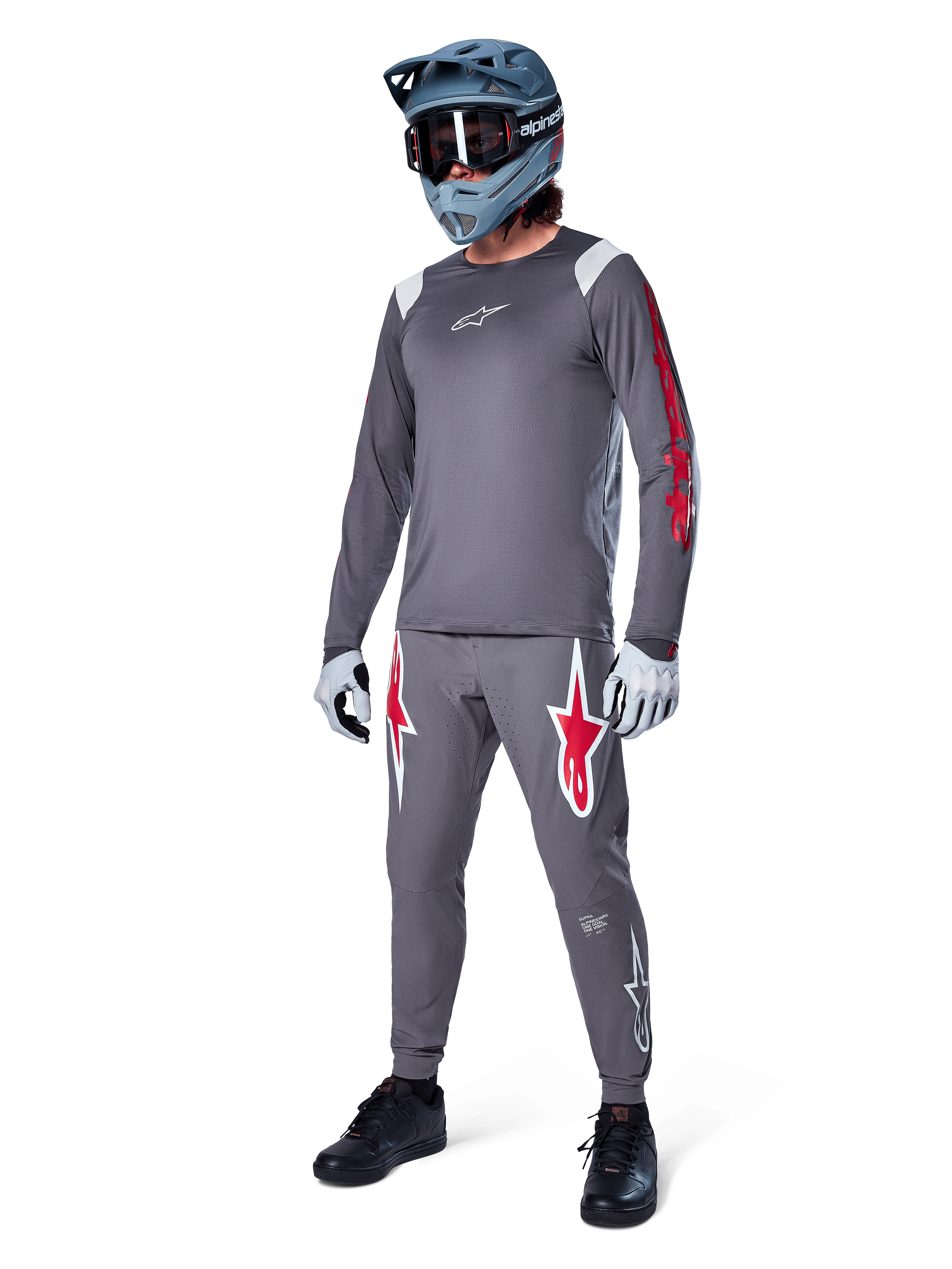 Camisola de Manga Comprida Alpinestars A-Supra Astar em cinzento escuro, com branding vermelho nas mangas e logótipo branco no peito, usada por um ciclista com calças cinzentas a condizer, capacete integral cinza-azulado e luvas de performance, equipamento coordenado de BTT para gravity