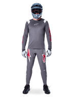 Ciclista de MTB com camisola de manga comprida Alpinestars A-Supra Astar, cinzento escuro com pormenores vermelhos e logótipo branco no peito, calças cinzentas a condizer com grandes grafismos de estrelas vermelhas, capacete integral azul com óculos, luvas cinza claro e sapatos pretos, conjunto de equipamento profissional de mountain bike