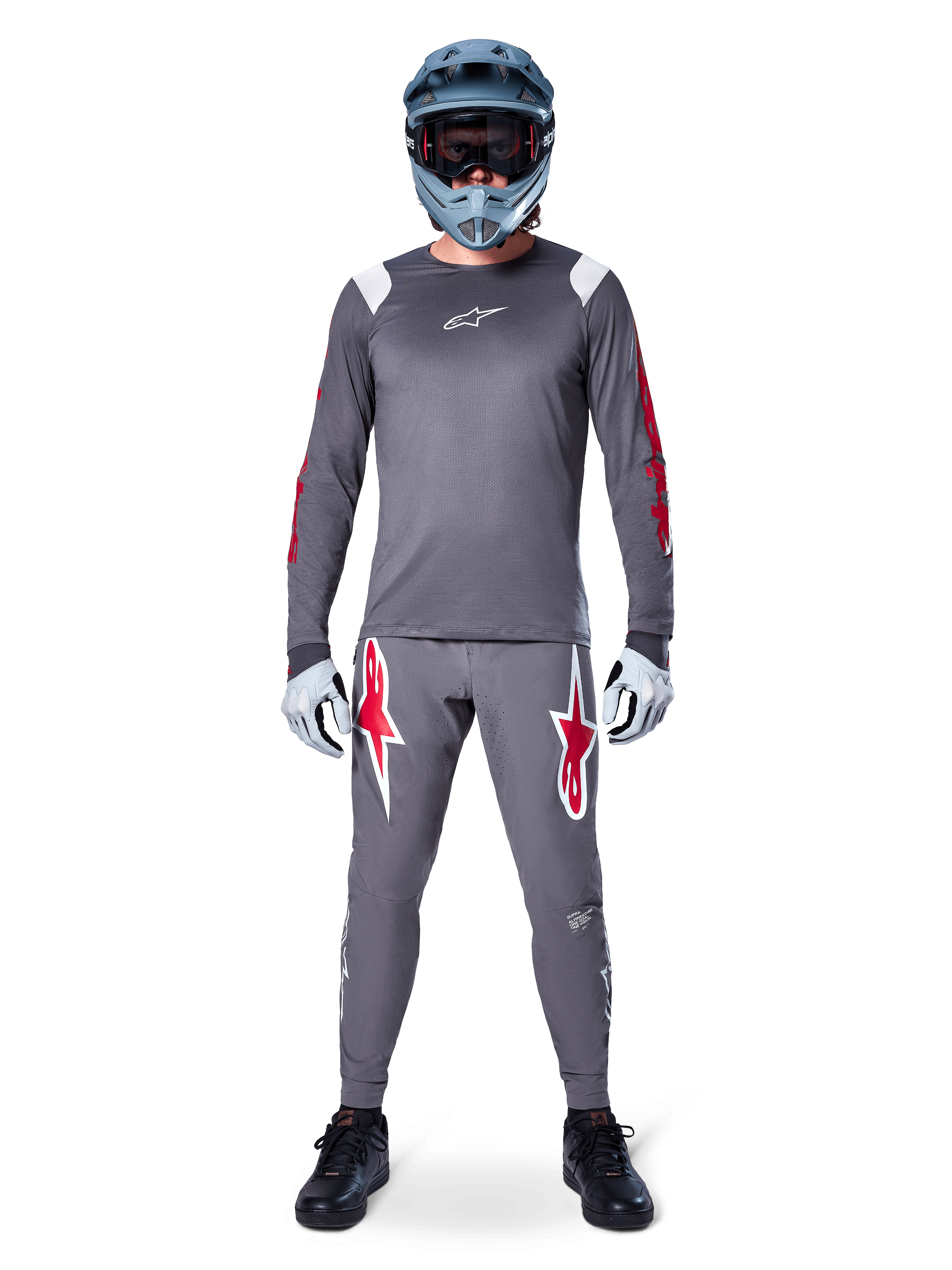Ciclista de MTB com camisola de manga comprida Alpinestars A-Supra Astar, cinzento escuro com pormenores vermelhos e logótipo branco no peito, calças cinzentas a condizer com grandes grafismos de estrelas vermelhas, capacete integral azul com óculos, luvas cinza claro e sapatos pretos, conjunto de equipamento profissional de mountain bike