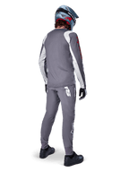 Camisola Alpinestars A-Supra Astar, camisola de BTT de manga comprida, cinzento escuro com detalhes brancos nas mangas e logótipo vermelho, vista traseira de ciclista a usar calças cinzentas a condizer e capacete integral azul, vestuário de ciclismo de performance