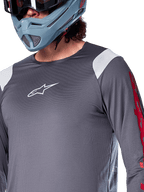Camisola de Manga Comprida Alpinestars A-Supra Astar, cinza escuro com painéis brancos nos ombros e grafismos vermelhos na manga, usada com um capacete integral cinza-azulado claro, vestuário de BTT de alta performance