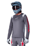 Alpinestars A-Supra Astar Jersey de manga comprida, jersey de BTT, cinzento escuro com detalhes em vermelho e branco, ajuste de performance, usado com calças cinzentas a condizer com gráficos de estrelas vermelhas e brancas, equipamento completo de ciclismo off-road com capacete integral e luvas