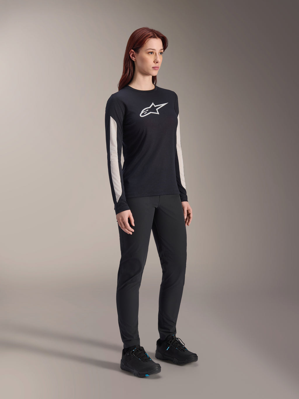 Mulher a usar equipamento de MTB Alpinestars, camisola Stella A-Dura Elite preta com logótipo branco no peito e painéis nas mangas, calças Stella A-Dura pretas, sapatos de ciclismo pretos com detalhes azuis, conjunto de performance completo para mountain bike
