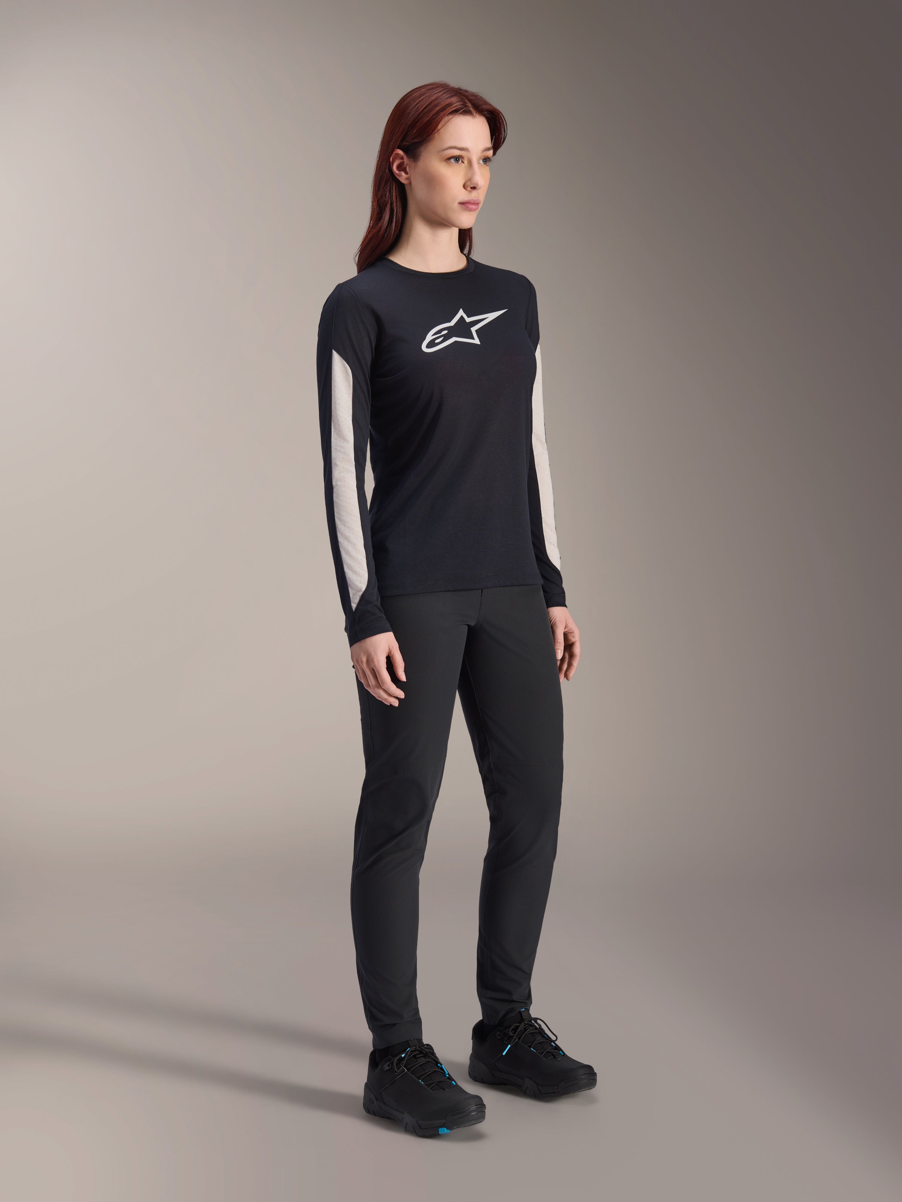 Mulher a usar equipamento de MTB Alpinestars, camisola Stella A-Dura Elite preta com logótipo branco no peito e painéis nas mangas, calças Stella A-Dura pretas, sapatos de ciclismo pretos com detalhes azuis, conjunto de performance completo para mountain bike