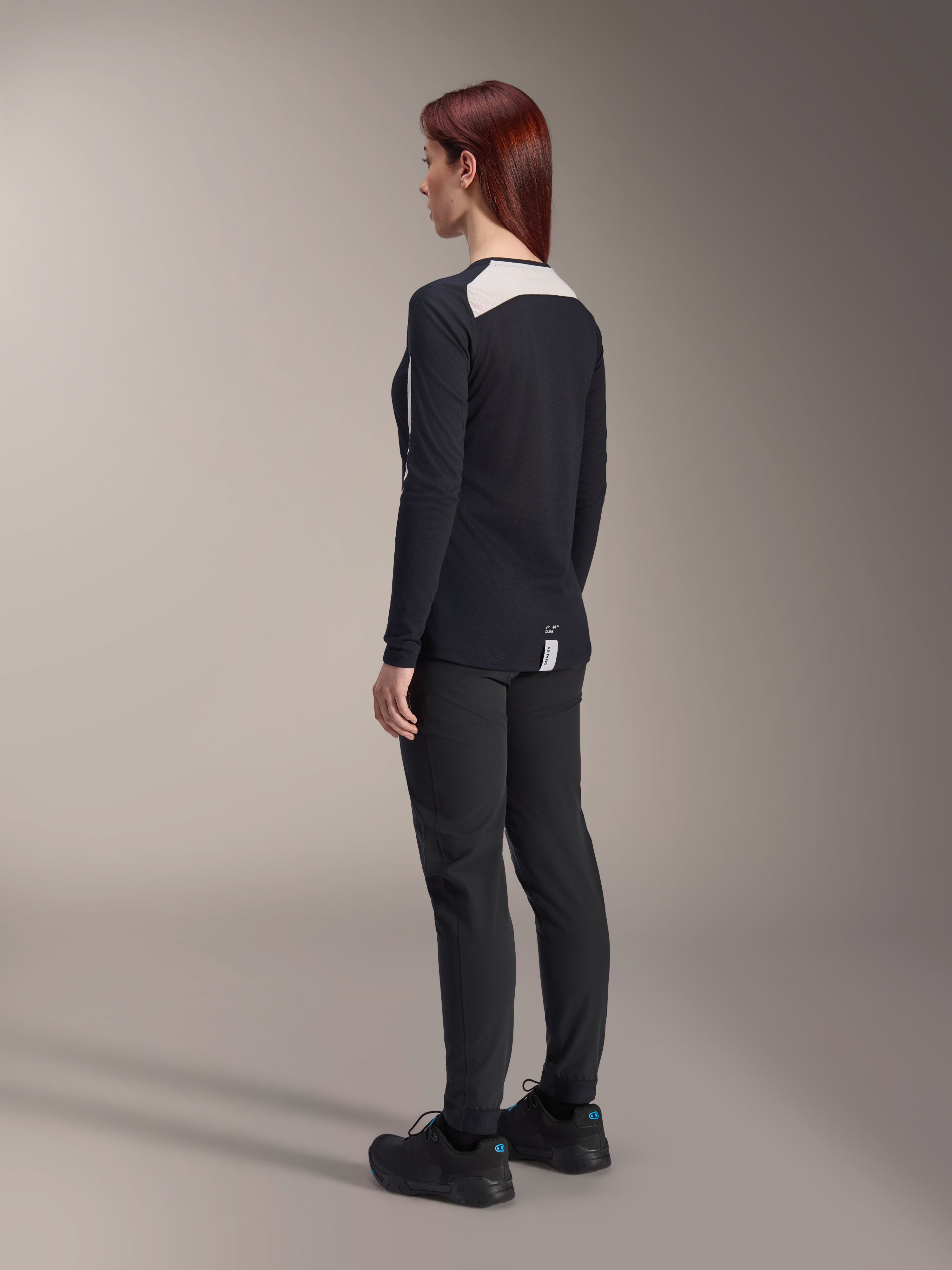 Ciclista feminina a usar calças Alpinestars Stella A-Dura e jersey Stella A-Dura Elite, pretos, vista traseira exibindo painéis de malha brancos nos ombros do jersey de manga comprida, calças técnicas slim-fit, equipamento de performance coordenado para mountain bike.