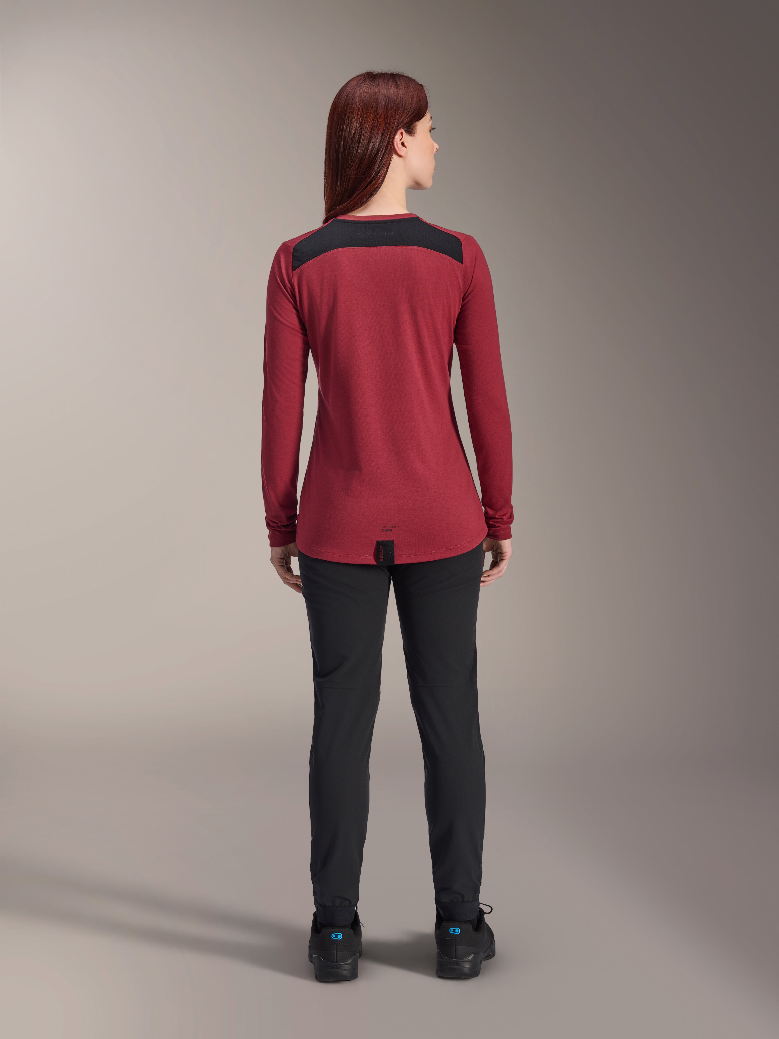 Alpinestars Stella A-Dura Dri Astar Jersey de Manga Comprida para Mulher, vermelho borgonha com painéis de malha pretos nos ombros, vestuário técnico de MTB visto de costas com calças pretas, design de cauda longa para maior cobertura ao pedalar
